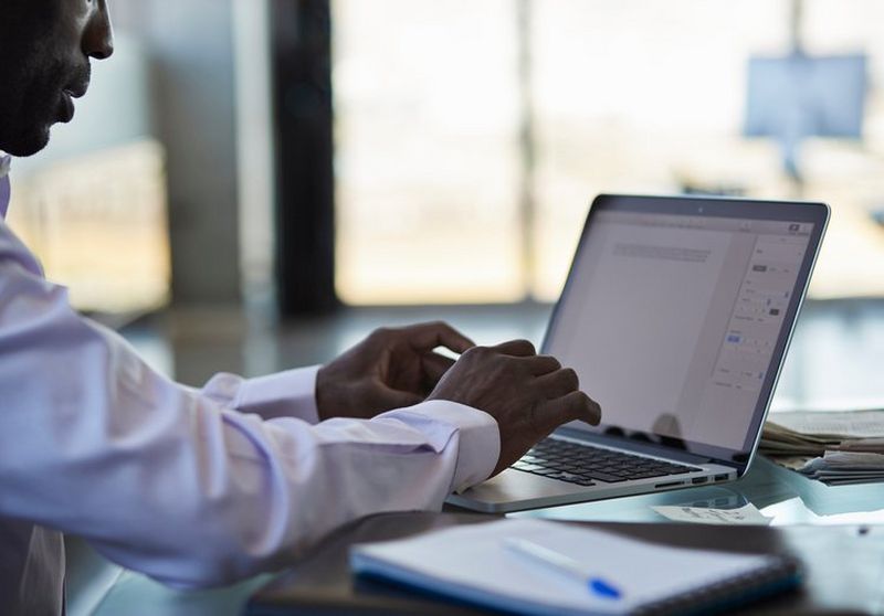 Close-up shot of a man in a crisp white shirt typing into a document on a laptop, with papers piled behind the computer.