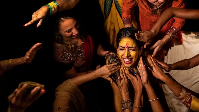 A South Asian woman stands centre shot. She is surrounded by many hands touching her shoulders and head in celebration