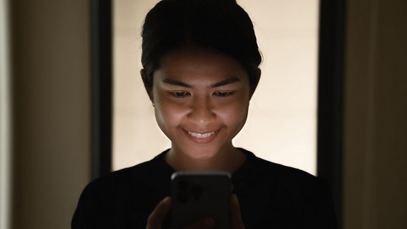 A smiling woman in a darkened room looks at her phone and the light from the screen gently illuminates her face.