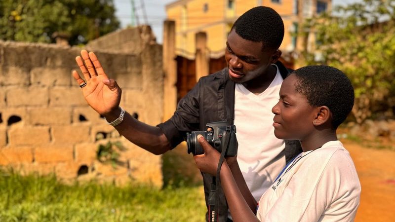 A young man holds up his hand, directing a young woman as she holds her camera. In the background are grass, trees and buildings.