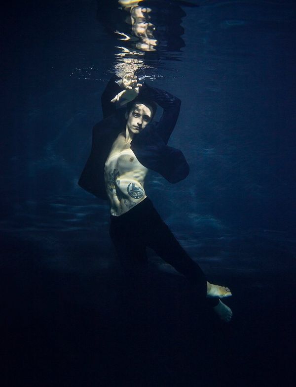 Sergei Polunin strikes a ballet pose underwater dressed in a suit, his chest exposed in the unbuttoned jacket. His upper body is reflected in the water above him.