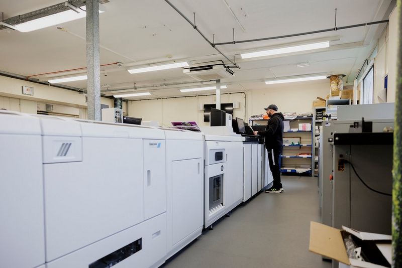 Een operator die de Canon imagePRESS-printer bedient in een goed verlichte werkruimte van de Universiteit van Loughborough. De omgeving bevat printapparatuur en goed georganiseerde rekken met benodigdheden