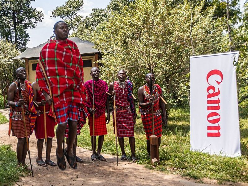 Ten men and women wearing the traditional dress of the Maasai tribe stand on a grassy verge next to a pop-up banner displaying the Canon logo. The man in the centre of the image jumps high into the air. © Peter Ndungu