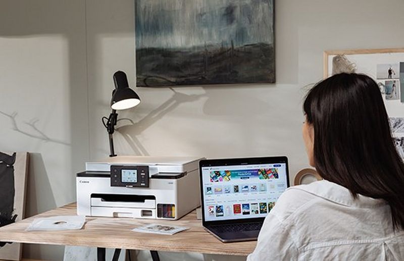 A woman sits working at a laptop, with a Canon MegaTank printer on the desk next to her.