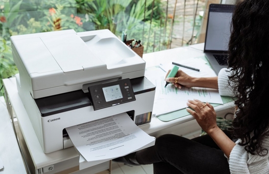 A woman sits at a desk using a highlighter on a printout. Next to her is a Canon MAXIFY printer, and a garden can be seen through the large window in front of her.