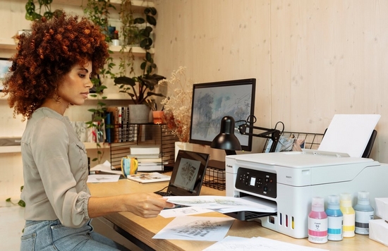 A woman sits at a home office desk on which is a laptop, a monitor and a Canon MegaTank printer plus four ink bottles.