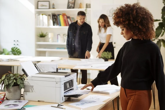 A woman picking up a printout from a Canon MAXIFY printer in an office with two other people in the background.  