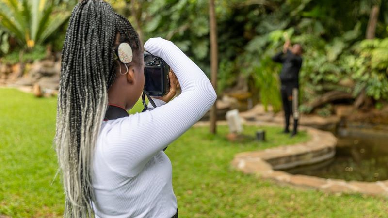 A young woman with a long ponytail holds a camera to her face, photographing a blurred figure in the distance.