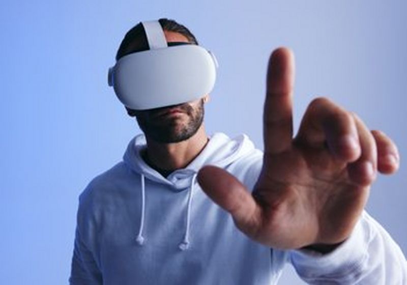 The upper body of a bearded man who is wearing a white hooded sweater and white VR headset. He has his left hand raised with finger pointed as though to press a button. He stands against a blue/grey gradient background.