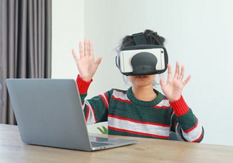 A child is sat at a table in front of a laptop. They are wearing a red, white and green striped sweater and a VR headset. They have both their hands raised in front of them, palms flat outwards, as though touching something.