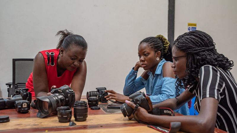 A woman holding a Canon camera leans on a table to show the back screen to two young women, who look at it intently.