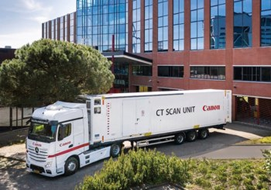A Canon branded lorry parks a mobile CT scanning unit, with CT SCAN UNIT and the Canon logo printed on the side. It is outside what appears to be a brown brick office building, surrounded by trees and bushes.