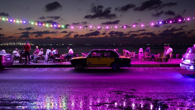 Groepjes mensen die bij elkaar zitten met uitzicht op een strand, geparkeerde auto's langs de weg en reflecterende paarse feestverlichting erboven. Gemaakt met Canon.