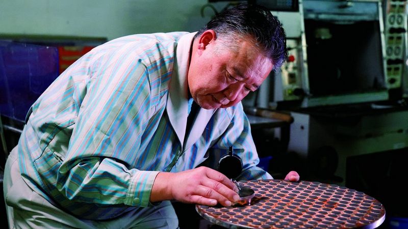 Een Japanse man in een gestreept blauw overhemd met een metalen stuk gereedschap in zijn hand leunt over een grote metalen schijf. Hij lijkt in een fabriek te zijn.