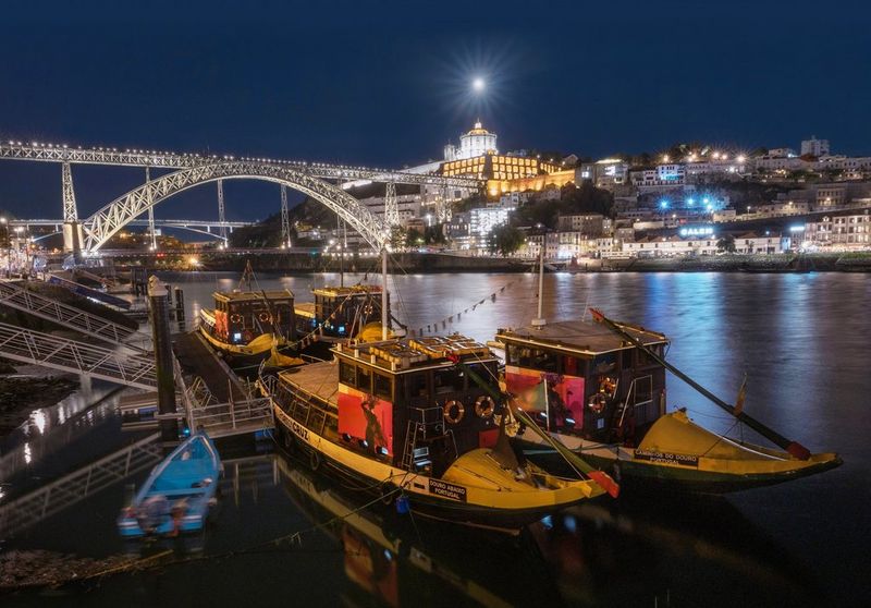 La Luna brilla nel cielo notturno, sopra un fiume con ponte, barche e un paesaggio urbano alle spalle. Scatto realizzato con Canon EOS R8.
