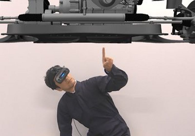 A man dressed in black is wearing a black Canon MREAL X1 mixed reality headset that covers his eyes. It has a neon blue stripe in the space between where his eyes should be. He crouches beneath a virtual representation of the chassis of a vehicle and points a finger to a spot just off the centre.   