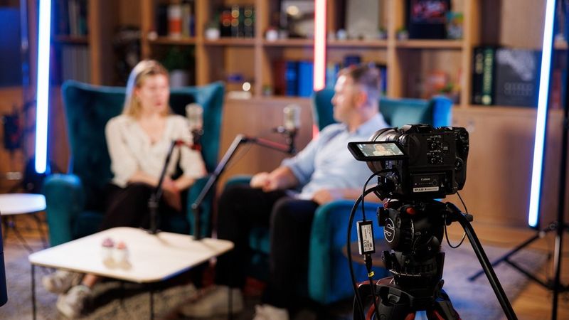 Two blurred people in armchairs, talking to each other. In the forefront of the image is a Canon camera on a tripod, filming them as they talk.