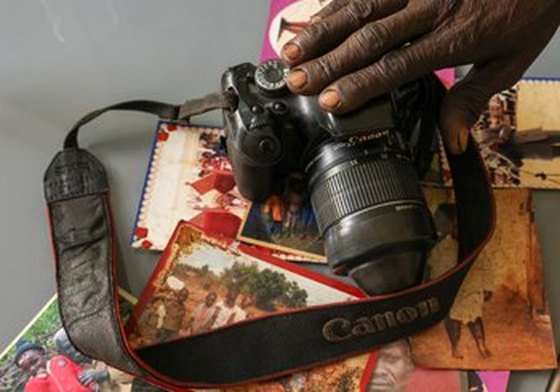 Some old photographs are spread out on a table, with a Canon camera sat on top. A hand reaches to touch the top of the camera.
