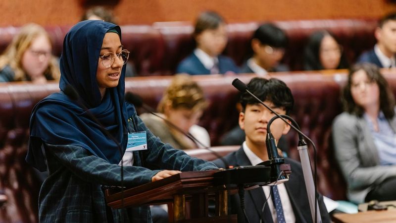 A young woman in glasses and a dark blue headscarf stands at a lectern, speaking into microphones. Behind her are the high-backed leather benches of parliament, filled with other young people