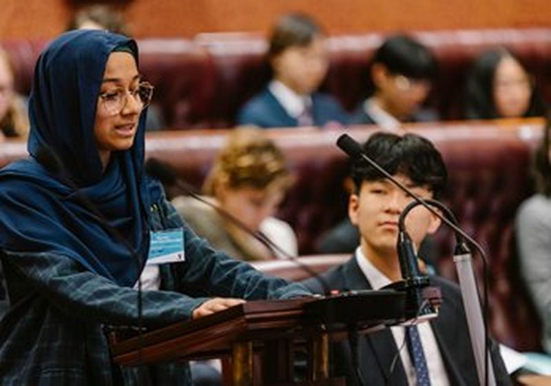 A young woman in glasses and a dark blue headscarf stands at a lectern, speaking into microphones. Behind her are the high-backed leather benches of parliament, filled with other young people