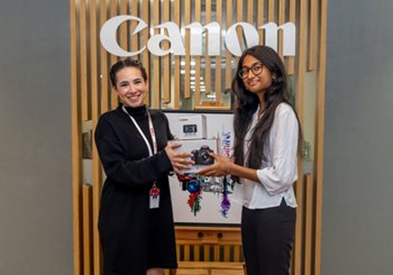 Two young women stand in front of a Canon sign. One passes a boxed camera to the other.
