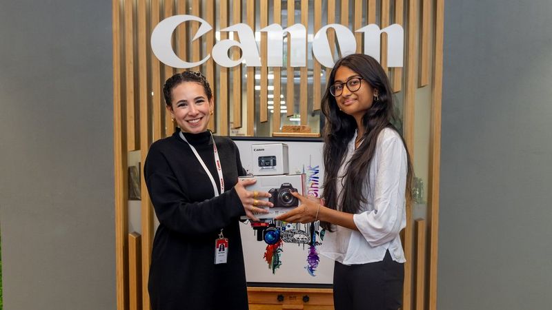 Two young women stand in front of a Canon sign. One passes a boxed camera to the other.