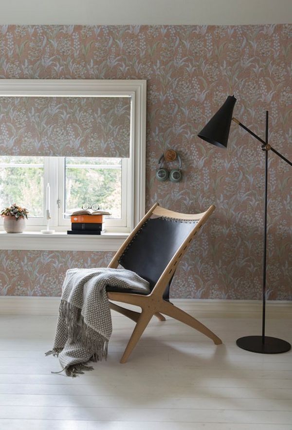A chair, with clothing draped across it, and a simple downward facing floor lamp, set against the reproduction wallpaper with Lily of the Valley pattern.