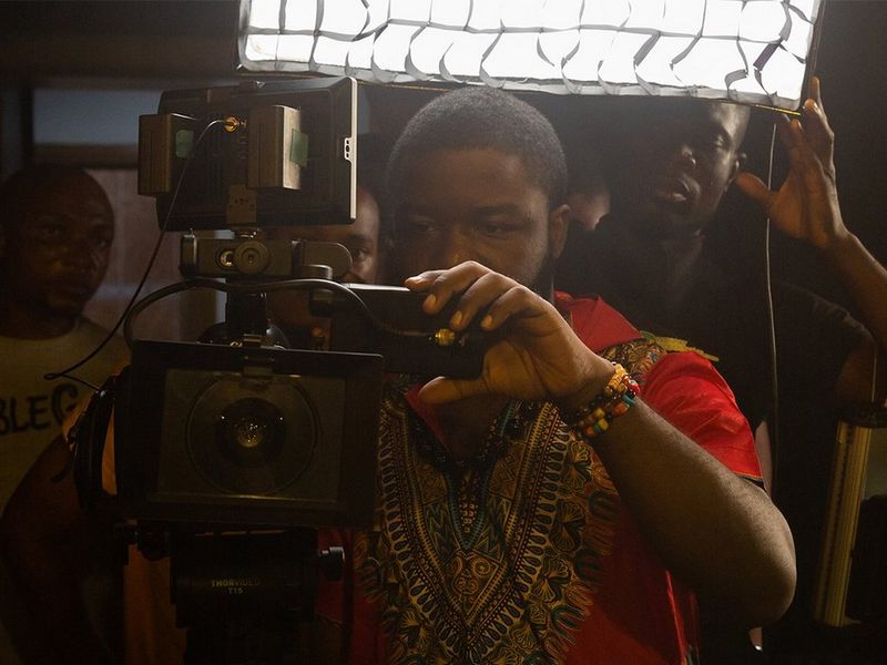 Um homem com uma t-shirt vermelha está a segurar e a olhar através de uma câmara Canon em Lagos, Nigéria. Atrás dele, dois homens olham na mesma direção.