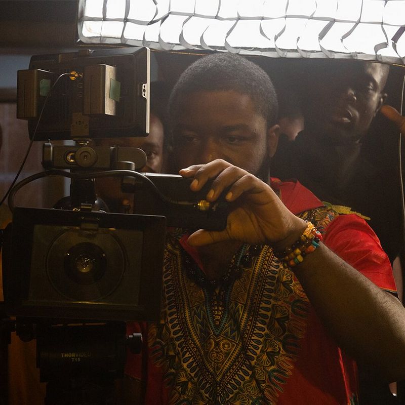 A man wearing a red t-shirt is holding and looking through a Canon camera in Lagos, Nigeria. Behind him, two men look in the same direction.