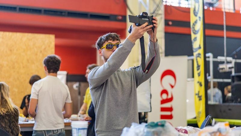 A young man in yellow sunglasses holds up a Canon camera to take a selfie. Behind him are other students and a banner of the Canon logo.