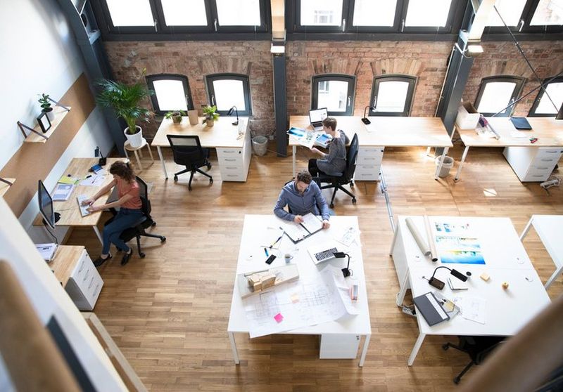 Wide overhead shot of a creative office layout with people working at desks adorned with computers, lamps and brochures.