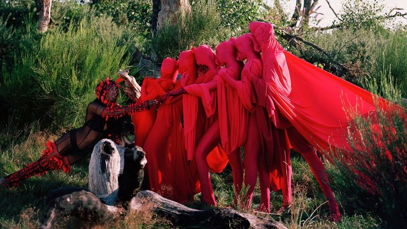 Six upright bodies, shrouded in red sheets from head to toe, cling on to each other, pulling away from a figure on the right, which is near naked, except for underwear, red stockings, sleeves and a headdress. 