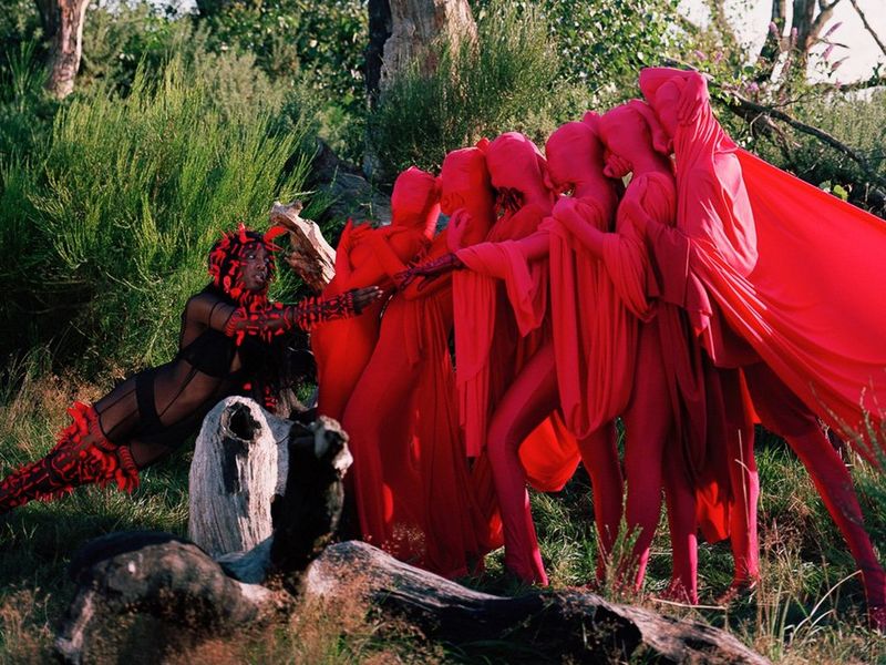 Six upright bodies, shrouded in red sheets from head to toe, cling on to each other, pulling away from a figure on the right, which is near naked, except for underwear, red stockings, sleeves and a headdress.