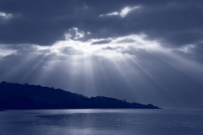 Un paysage marin monochrome aux teintes bleues, où les rayons du soleil traversent les nuages sombres au-dessus de la silhouette d'un littoral vallonné.