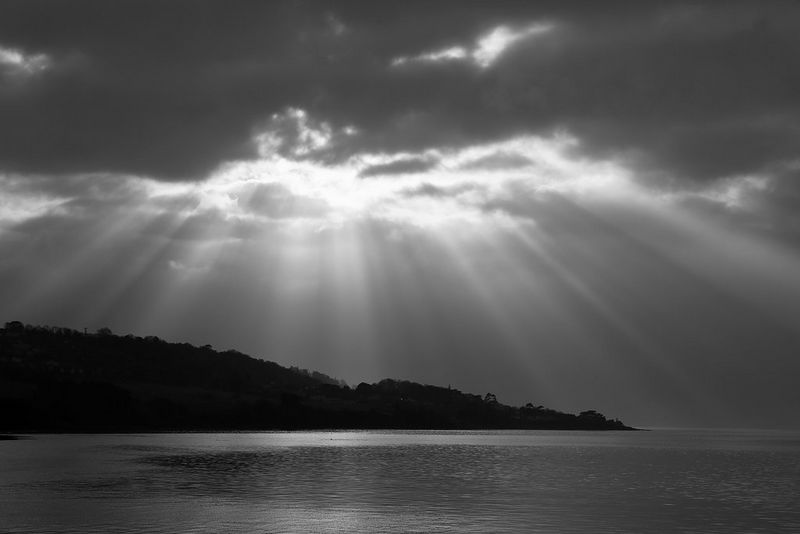 A black and white seascape, with the sun's rays shining through dark clouds above the silhouette of a hilly coastline.