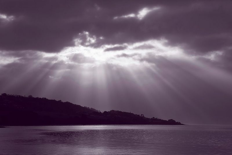 A purple-tinted monochrome seascape, with the sun's rays shining through dark clouds above the silhouette of a hilly coastline.