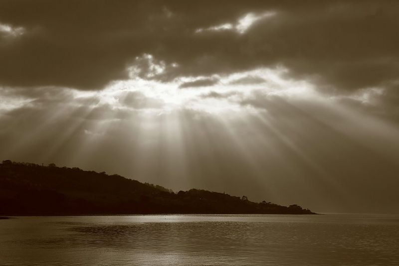 A sepia-tinted monochrome seascape, with the sun's rays shining through dark clouds above the silhouette of a hilly coastline.