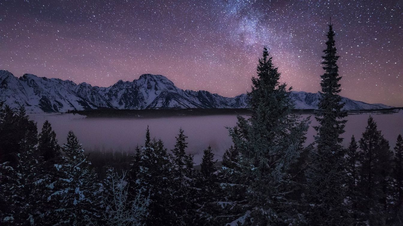 Uma imagem de paisagem de pinheiros, um lago e montanhas cobertas de neve sob um céu estrelado.