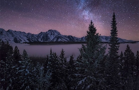 A landscape image of pine trees, a lake and snow-dusted mountains under a starry sky.
