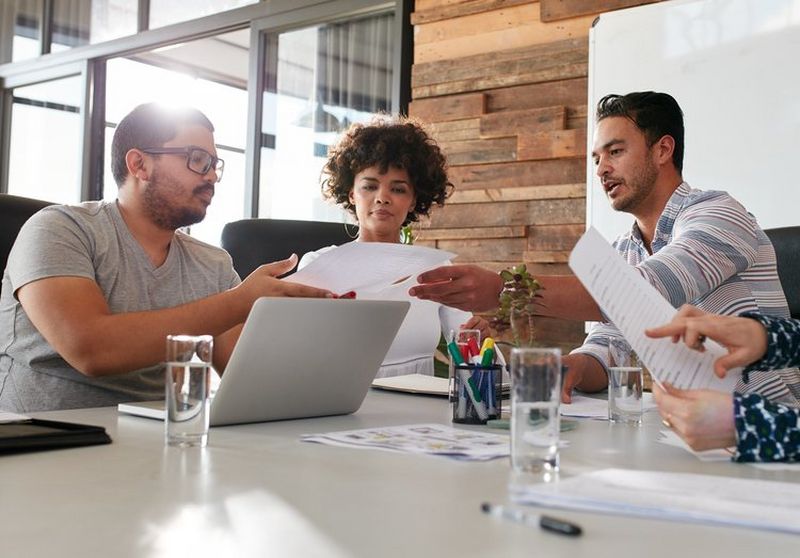 Two men and a woman pass papers over a large white desk, with open laptop, pens in a holder and glasses of water.