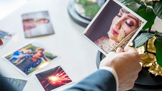 A hand holding a square image of a bride's face with traditional jewellery and clothes. Other pictures seen in the background spread over the table. 