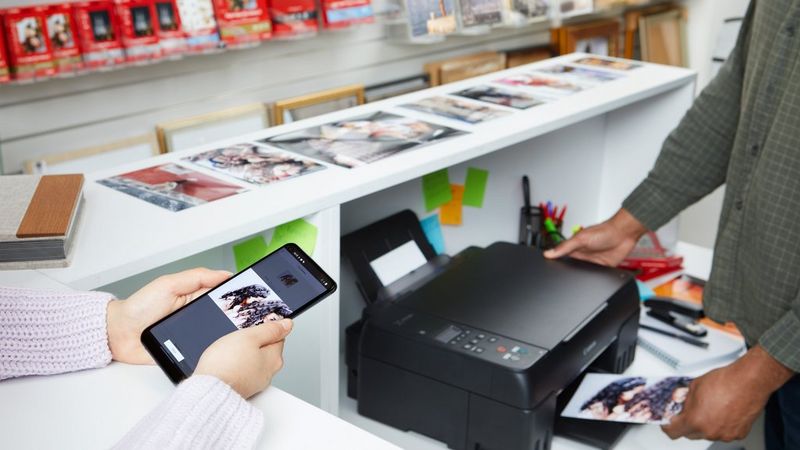 Планшет, на котором открыто приложение Canon PRINT, лежит на принтере Canon, из которого появляется фотография; рядом на столе можно увидеть еще несколько фотокарточек.