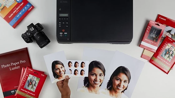 A Canon printer on a white desk with Canon camera, photo paper of different sizes spread around it and a hand holding passport-size and other size pictures of the same woman.
