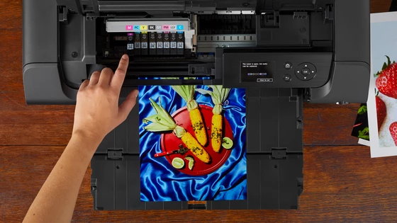 O imprimantă Canon deschisă din partea de sus, cu cartuşele de cerneală vizibile şi o mână arătând spre unul dintre ele. O copie imprimată a unei fotografii colorate cu alimente şi alte imagini lângă aceasta.