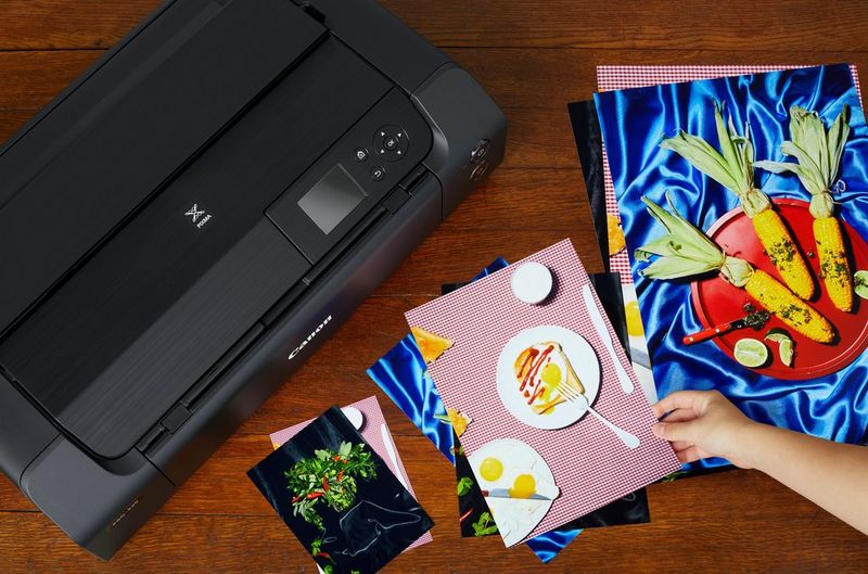 A woman's hand holds one of a number of borderless prints of different sizes, on a tabletop next to a Canon PIXMA PRO-200 printer.