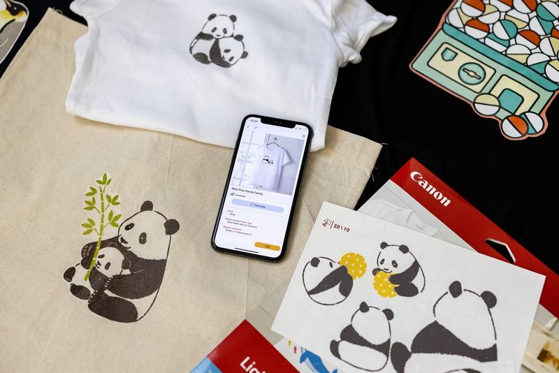 Слика снимена одгоре прикажува слики од панди на телефон, отпечатени на хартија за пренос на слики, како и на торба со крем-боја и бебешко боди.