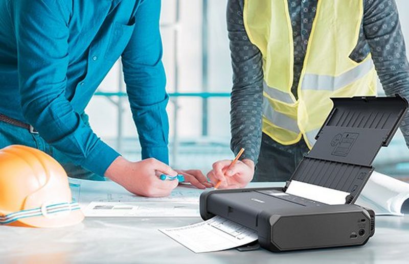 Two construction workers printing off documents on a Canon PIXMA TR150. An orange hard hat is on the table beside them.