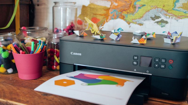 Een Canon-printer op een houten bureau print een kleurrijke knutselsjabloon. Het bureau staat vol met knutselbenodigdheden, waaronder potten met pompons en stiften. Origamifiguren liggen verspreid over de printer. Op de achtergrond hangt een kaart van Europa aan de muur.