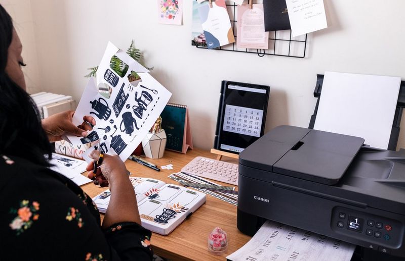 A woman cuts out Creative Park templates while sitting at a wooden desk, on which sits a Canon PIXMA printer and a selection of crafting materials.