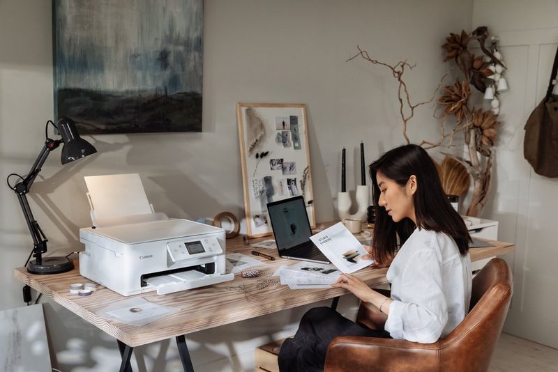 A woman looks at a pile of papers, a Canon PIXMA printer and other stationery sitting on the table. 